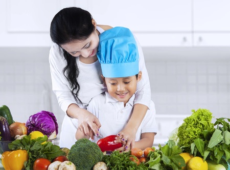 asian mother is teaching her son to cut vegetable at home